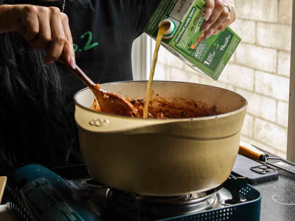 A person pours vegetable broth from a carton into a beige pot filled with a red sauce, stirring with a wooden spoon on a stove.